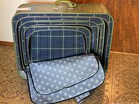 Four green canvas-sided luggage cases nested inside each other, showing plaid interior of each case and a protective blue patterned cover inside smallest case.