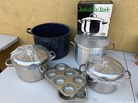 Photo of two steel covered cooking pots with lids, a large blue enamel steel pot with speckled pattern and one handle missing, an aluminum covered casserole, and two muffin pans on a table.