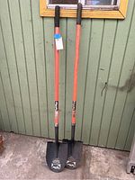 Front view of two shovels leaning against green siding, showing orange fiberglass handles with black grips and black metal blades