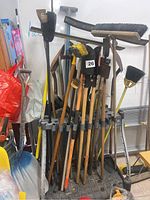 Photo showing a large assortment of garden tools leaning against a wall, including brooms, rakes, shovels, hoes, and other long-handled tools, primarily wooden handles with metal heads.
