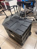 Closed state of black collapsible rolling bucket showing handle and wheels, placed on tiled floor near office chairs.