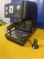 Front and side views of the black Saeco Via Venezia espresso machine showing control buttons, portafilter, steam wand, and tamper on yellow and blue background.