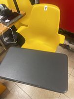 Yellow plastic chair with armrests, showing scratches and wear. Attached is a large black rectangular tray mounted on a swiveling arm. The base has a perforated circular footrest on caster wheels.