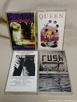 Four cassette tapes including Temple of the Dog, Queen, Rolling Stones, Rush shown in plastic cases on fabric surface.