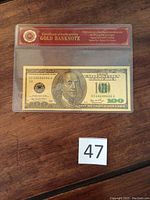 Sealed gold foil $100 banknote with visible Certificate of Authenticity in protective sleeve.