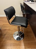 Pair of black vinyl padded bar stools with vertical stitching, adjustable height chrome pedestal bases with footrests, placed on wooden floor beside counter.