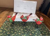 Three cardinal bird figurines wearing green scarves standing on black metal legs on green holly berry Christmas fabric. White styrofoam packaging visible behind the birds.