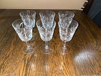Six vintage crystal stemmed glasses grouped on a wooden table showing the etched floral/starburst pattern around the bowl and tall slender stems.