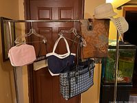 Four bags hanging from hangers on metal rack, two straw hats on right shoulder of rack, brown patterned scarf and pair of sunglasses also visible.