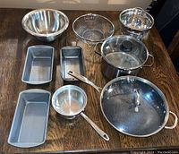 Full view of all cookware items on the table showing strainers, mixing bowl, loaf pans, pots and pans.