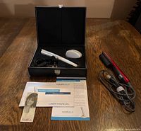 Wide shot showing HairMax LaserComb, charger, base, user manual in black case, red and black hair straightening tool with power cord on wooden table.