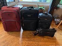 Four suitcases and one travel bag arranged in a line on wooden floor by a window, showing size and color variation.