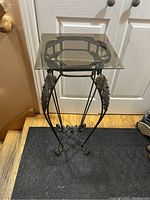 Full view of metal side table with glass top, standing on a black mat in front of a white door. Shows decorative leaf metalwork and curled feet.
