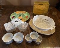 Mixed kitchen items on wooden table: colorful oval serving dish with fruit designs, oval serving tray, gold metal tin, plastic colander basket, square plastic containers, and white ceramic ramekins.