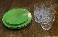 Five green plastic plates (3 large, 2 medium) stacked beside five clear plastic tumblers on wood floor.