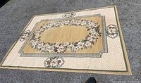 Full outdoor view of the large beige floral rug laid flat on gravel surface showing overall pattern and size.