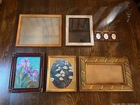 Top-down photo showing two framed floral paintings and five empty frames arranged on a wooden floor.