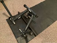 Full view of the black tubular metal frame bike trainer on a black rubber mat, showing its foldable legs and frame shape