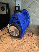 Blue Campbell Hausfeld cordless inflator pump powered via cord plugged in outlet on kitchen counter, with LED indicators lit, flashlight facing front.