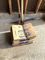 Stack of folded, bundled eco-friendly brown paper lawn bags on concrete ground with parts of two pick axes behind.