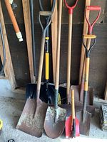 Photo showing six shovels and scoops leaned against a wall, displaying variety in blade style and handle design.