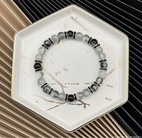 Photo shows a 7-inch black and grey crackled glass bead bracelet with silver spacers, placed on a hexagonal white dish with gold accents.