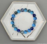 Top view of the blue beaded bracelet showing round alternating blue solid and cracked glass beads on a hexagonal white dish with gold trim.
