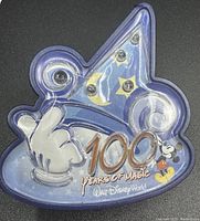 Photo showing Disney 100 Years of Magic light up collector pin featuring Mickey Mouse's hand holding a wand, stars and moon decorations, and logo text.