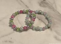 Photo shows two elastic beaded bracelets with crackled glass beads in pastel colors and silver-tone sparkle accent beads arranged overlapping on a marble surface.