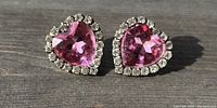 Close-up of a pair of pink heart shaped earrings set with clear round stones around the edge, photographed on a wooden surface with natural light.