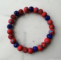 Photo of the full 7" wrist bracelet showing red beads with blue splatter and alternating red and blue rhinestone encrusted beads on a neutral background.