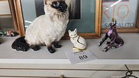 Three cat figurines on a shelf: ceramic Siamese, porcelain trinket box cat with gold trim and rhinestones, purple wooden cat.