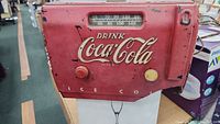Front view of Coca-Cola radio with 'DRINK Coca-Cola' branding, tuning dial, and damaged lower right front corner showing 'ICE COL' text