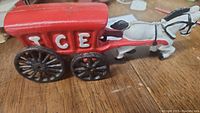 Side view of cast iron horse-drawn red ICE wagon toy with black wheels on wooden surface.