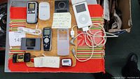 Collection of electronic devices on a red and orange woven mat, clearly showing various items including iPod, Olympus digital voice recorder, Belkin photo viewer, FM transmitter iTrip, travel alarm clocks and cables.