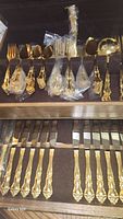 Full set of goldware flatware including knives, forks, spoons, and serving utensils inside dark lined wood case with some pieces still wrapped in plastic sleeves.