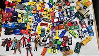 Wide view of multiple toy cars and action figures scattered on a white surface showing the variety of the collection