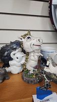 Front view of white ceramic bobble-head unicorn with gold horn, behind it a white porcelain mother and foal unicorn, on wooden surface.