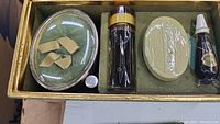 Top-down view of vintage Faberge Aphrodisia set in green velvet-lined presentation box showing sealed perfume spray, wrapped bar soap, small sealed bottle, and oval compact with green velvet insert.