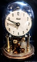 Vintage Kundo mantel clock under a glass dome. White dial with black Arabic numerals and black hands. Brass-toned base and pendulum balls beneath the dial.