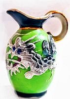 Front angled view of miniature ceramic pitcher showing green body, raised silver dragon with colorful accents, black rim with gold trim and handle.