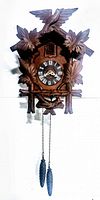 Front view of vintage hand carved wooden cuckoo clock showing carved leaves, bird, clock face with Roman numerals, and hanging pine cone weights.