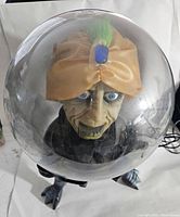 Front view of the Gemmy Halloween fortune teller inside a clear dome showing a tan turban with a blue gem and green feather, expressive face with light-up eyes.