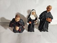Three religious-themed figurines arranged standing and kneeling on a white background showing frontal view with bishop kneeling, nun and priest standing with detailed facial expressions and attire.
