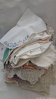 Pile of folded vintage linens and crochet doilies featuring embroidered floral designs.
