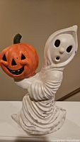 Full view of the ceramic ghost figurine holding an orange pumpkin with carved features.