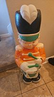Full front view of vintage blow mold drummer boy figure with large black hat and orange uniform standing on tile floor.