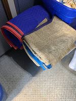 Photo showing one blue quilted packing blanket with red edging next to two burlap blankets with blue edging on a textured floor.