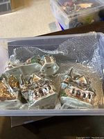 Four ceramic Christmas village decorative houses in a plastic container with bubble wrap, showing detailed paintwork including wreaths and garlands.