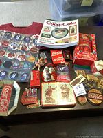 Overview of assorted Coca-Cola collectibles including Price Guide book, fridge magnets, ornaments, playing cards, mini bottles, T-shirt, and crate on table.